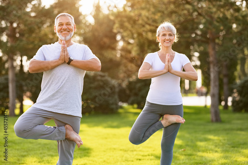 Positive senior couple performing yoga on green lawn in park during sunrise