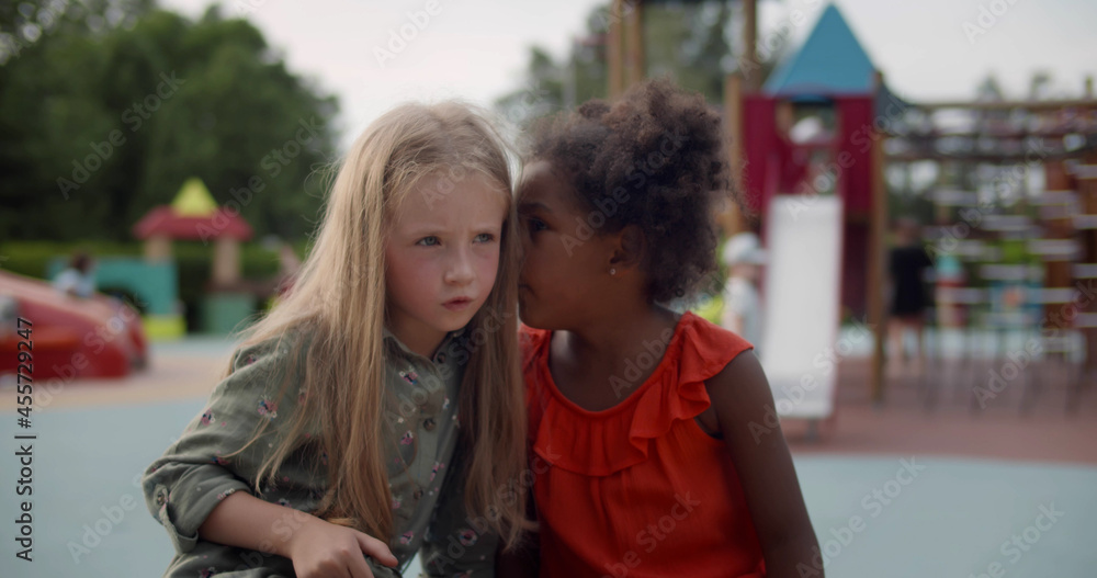 Medium shot of multiracial little children tell secrets on bench outdoors