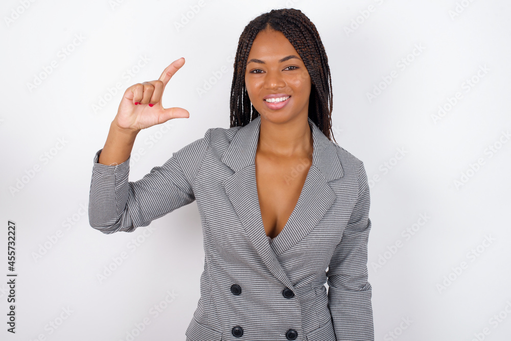 Young african american business woman with braids over white wall smiling and gesturing with hand small size, measure symbol. Young african american business woman with braids over white wall smiling and gesturing with hand small size, measure symbol.