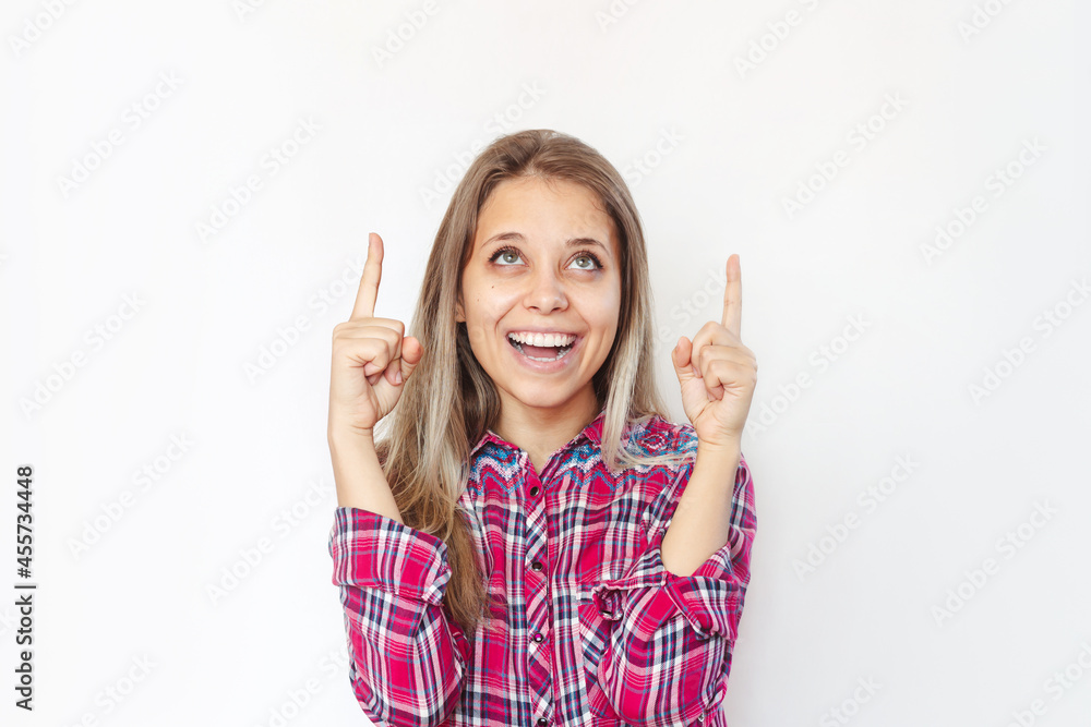 A young charming smiling blonde woman in the red plaid shirt points up to a copy empty blank space for text or design with two fingers presenting product isolated on a white background A young charming smiling blonde woman in the red plaid shirt points up to a copy empty blank space for text or design with two fingers presenting product isolated on a white background