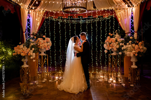 newlyweds at evening wedding ceremony in park among light bulbs and garlands.