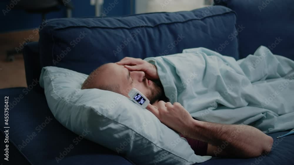 Close up of adult using oximeter for disease diagnosis. Medical device with decreased oxygen saturation on finger of sick person laying on couch. Man checking lower pulse pressure Close up of adult using oximeter for disease diagnosis. Medical device with decreased oxygen saturation on finger of sick person laying on couch. Man checking lower pulse pressure