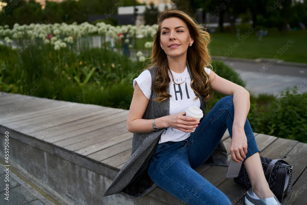Thoughtful female tourist in casual clothes holding coffee to go and smartphone technology, pondering woman with caffeiene beverage and cellular phone thinking about weekend leisure spending