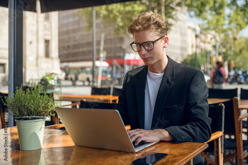 Billede på lærred A young businessman in a suit sits on the terrace of a cafe and works on a laptop