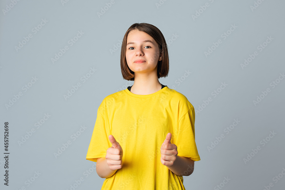 © Red Cristal - Portrait of beautiful cute teen girl in yellow casual tshirt looking at camera and showing thumbs up, standing over light grey background. Happy cute young girl showing okay