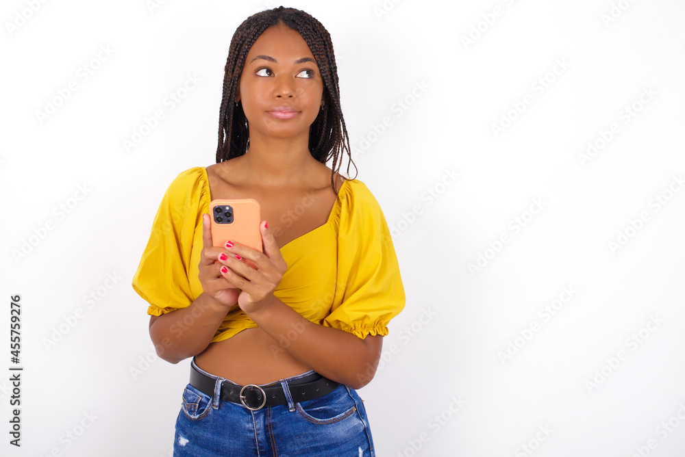 Portrait of Young african american woman with braids over white wall with dreamy look, thinking while holding smartphone. Tries to write up a message. Portrait of Young african american woman with braids over white wall with dreamy look, thinking while holding smartphone. Tries to write up a message.