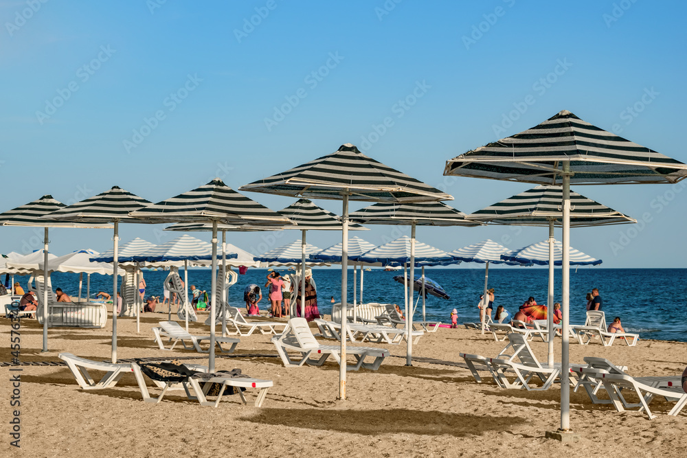 Zaliznyi Port, Ukraine - July 23, 2020: Striped umbrellas and white sun loungers on the beach at Zaliznyi Port (Kherson region). People rest and sunbathe on the Black Sea coast