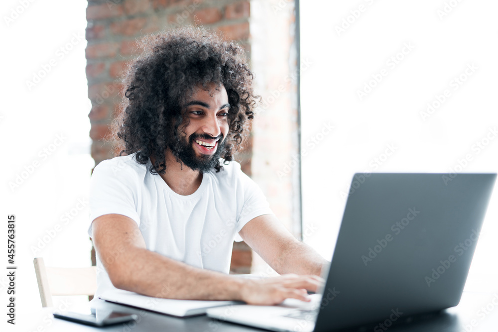 An African-American student with black curly hair, smiling broadly and sitting at his desk studying online An African-American student with black curly hair, smiling broadly and sitting at his desk studying online