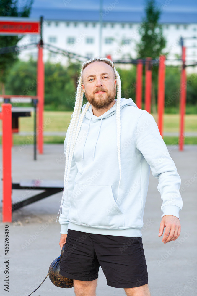 A man with an interesting braid hairstyle in a blue hoodie works out early in the morning on the street and lifts a kettlebell. A man with an interesting braid hairstyle in a blue hoodie works out early in the morning on the street and lifts a kettlebell.