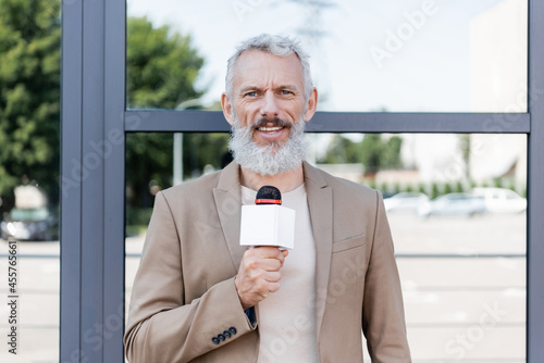 bearded announcer in blazer holding microphone and making reportage near building photo