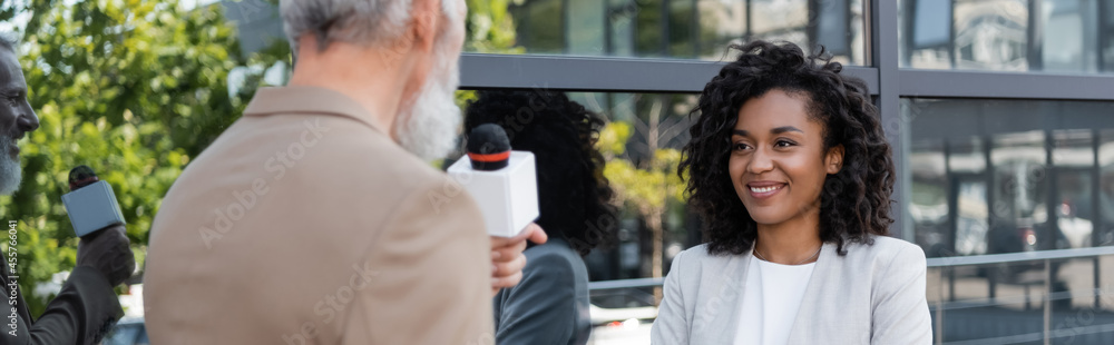 blurred and bearded reporter with microphone talking near smiling african american businesswoman, banner blurred and bearded reporter with microphone talking near smiling african american businesswoman, banner