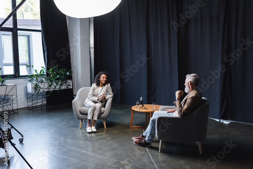 positive african american journalist in suit talking with businessman sitting in armchair during interview