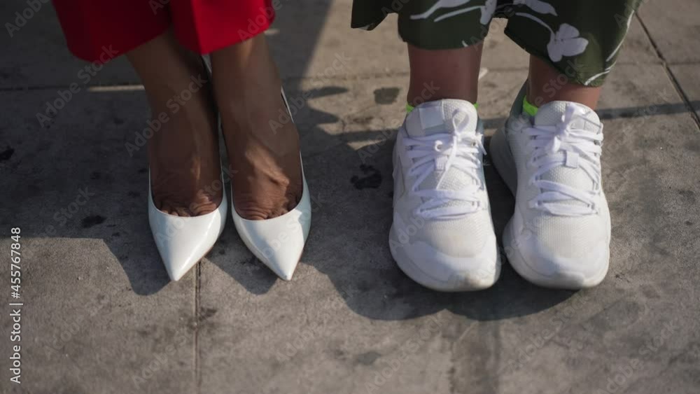 Close-up female feet in sneakers and slim legs in high-heels outdoors on pavement. Two unrecognizable Caucasian women sitting on bench in casual and formal outfit. Style and lifestyle concept Close-up female feet in sneakers and slim legs in high-heels outdoors on pavement. Two unrecognizable Caucasian women sitting on bench in casual and formal outfit. Style and lifestyle concept