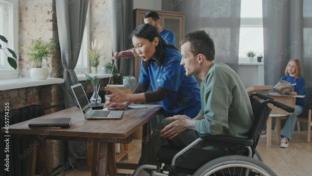 Tracking slowmo shot of multi-ethnic team of volunteers explaining young man with disability in wheelchair how to use internet on his computer while setting up wi-fi router in his apartment Tracking slowmo shot of multi-ethnic team of volunteers explaining young man with disability in wheelchair how to use internet on his computer while setting up wi-fi router in his apartment