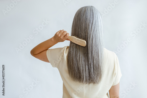 Lady in yellow blouse brushes straight silver hair standing on light grey background in studio backside view. Mature beauty lifestyle photo