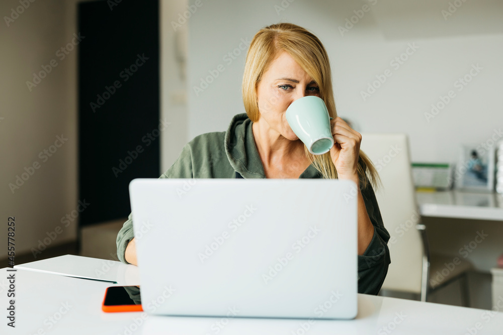 Middle age blonde woman using a laptop in a livingroom and drinking coffee on a mug Middle age blonde woman using a laptop in a livingroom and drinking coffee on a mug