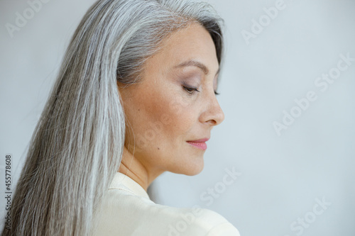 Pretty middle aged Asian woman with loose straight grey hair poses on light background in studio closeup side view. Mature beauty lifestyle photo