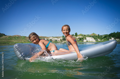 Two little cute girls in swimsuits swim on an inflatable mattress on a blue career lake. © bearok