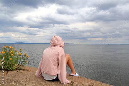 Female in a pink hood sweater looking out over the water. Sitting next to the flower tansy. Part of the Swedish lake called Vänern or Vanern. Lidköping, Sweden, Europe. photo