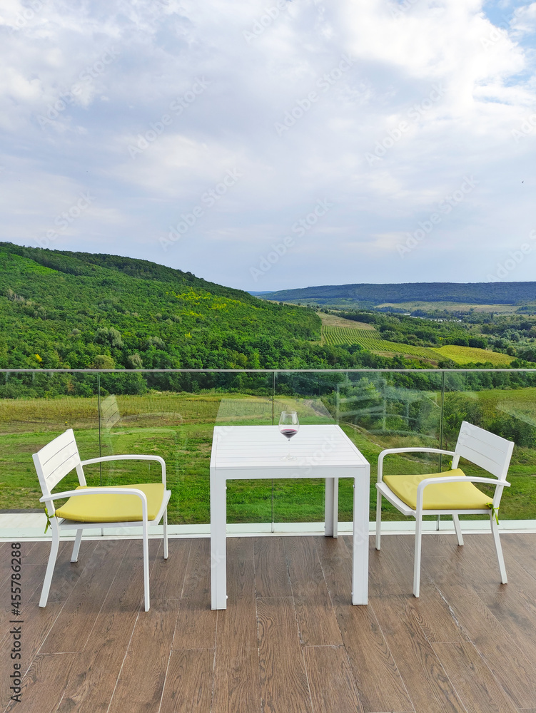 Table and two chairs with cushions on the empty balcony. White furniture on the terrace with a beautiful view of the forest in a hilly area. Background for recreational, relaxation and resting. Table and two chairs with cushions on the empty balcony. White furniture on the terrace with a beautiful view of the forest in a hilly area. Background for recreational, relaxation and resting.