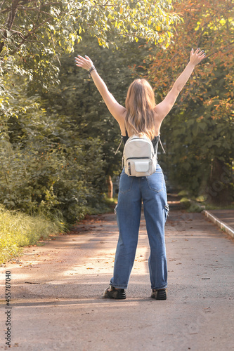 A young girl with a white backpack stands in full growth outdoors, two hands are raised up.