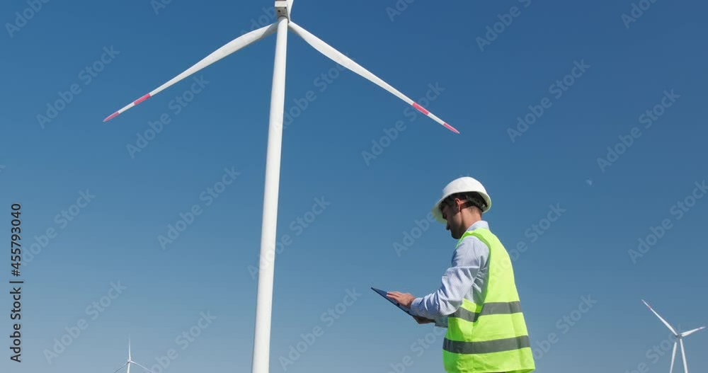 Mechanical engineer works with a tablet computer and looks at the rotation of the blades of a wind generator. Maintenance and tuning of the operation of wind turbines against the background. Mechanical engineer works with a tablet computer and looks at the rotation of the blades of a wind generator. Maintenance and tuning of the operation of wind turbines against the background.