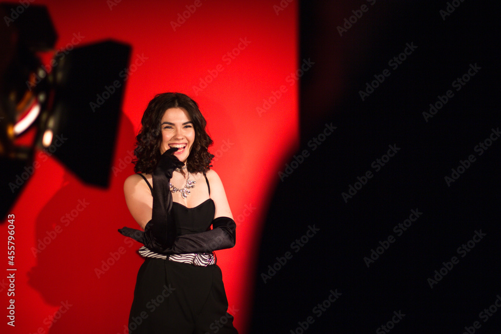 Young brunette girl in black overalls and black long gloves laughs posing for the camera on a red studio background. Fashion photo session