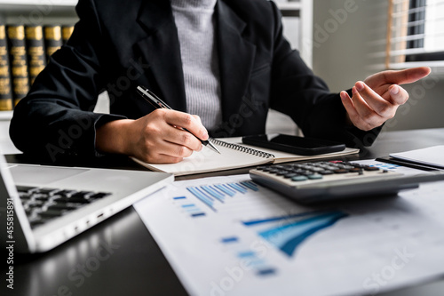 Close up of businesswoman or accountant writing in notebook while working analytic business report on a laptop computer at the workplace, investment, planning financial and accounting concept.