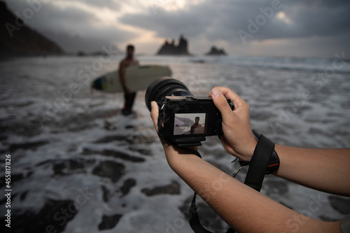 Close-up of the hands of a photographer holding his camera during a photo session with a surfer model at the beach during sunset - Photoshoot concept.