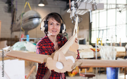 Portrait of happy female hobbyist in aviator headphones with unfinished monoplane model in aircraft workshop..