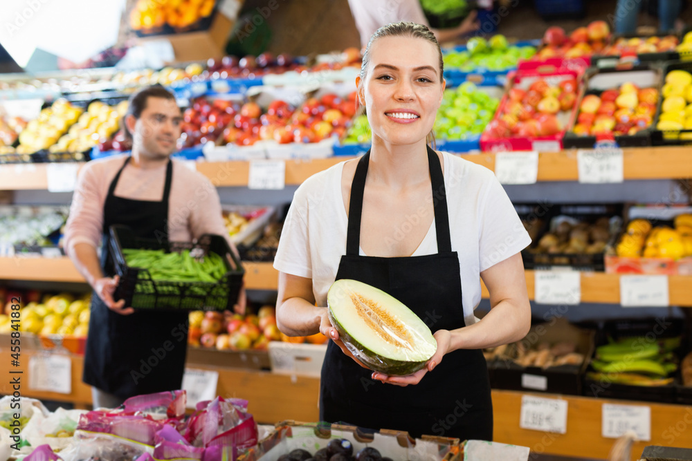 Young happy cheerful smiling female seller with half of melon and man on background having box of green beans on the supermarket