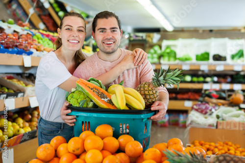 Glad family couple standing with full grocery cart after shopping in fruit store