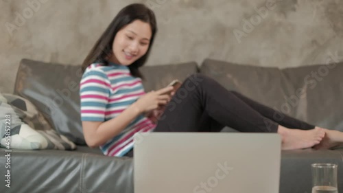 Beautiful young Asian woman working at home during quarantine lying down using mobile phone in living room while waiting for laptop chat call on internet e-commerce order, online shopping business. photo