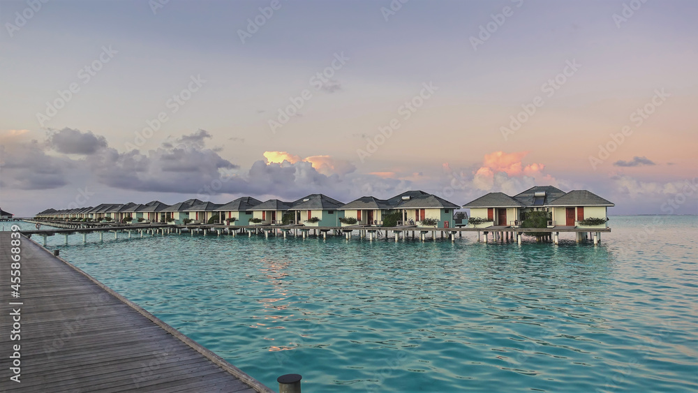 A calm morning in the Maldives. A wooden path and a row of water villas over aquamarine water. There are pink clouds in the blue sky. Delicate pastel shades