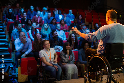 Speaker in wheelchair on stage talking to conference audience © KOTO