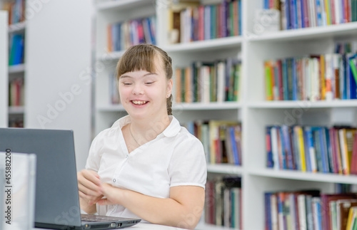 Smiling young girl with syndrome down uses a laptop at library. Education for disabled children concept