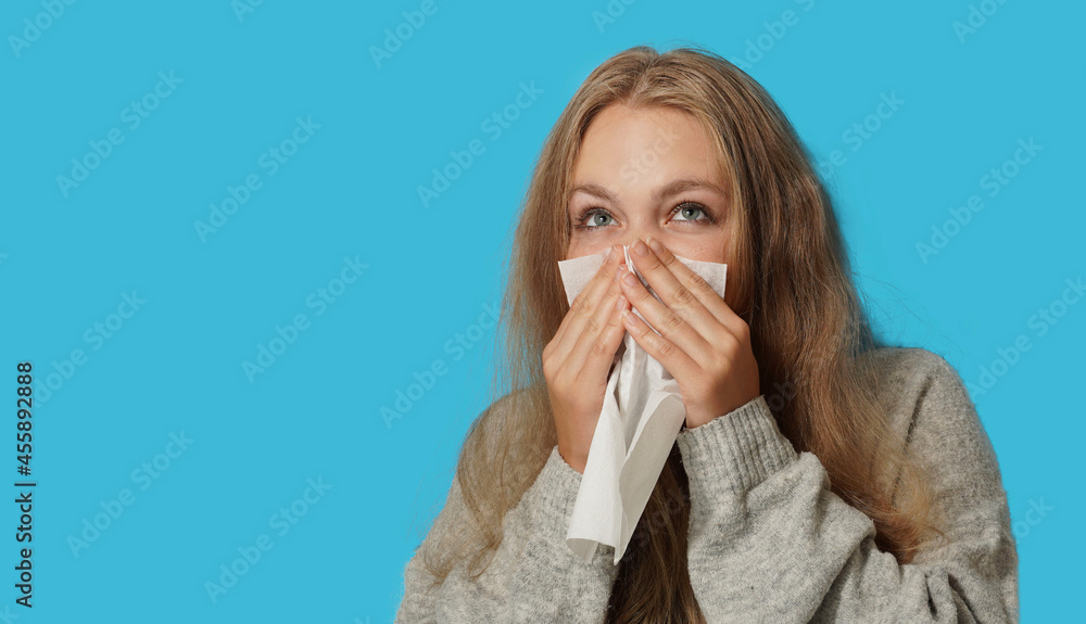 Young blond woman suffering from allergy on light blue background. Symptoms of cold or allergy. Woman sneezes in white tissue. Young blond woman suffering from allergy on light blue background. Symptoms of cold or allergy. Woman sneezes in white tissue.