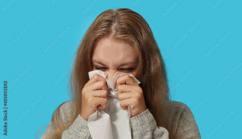 Young blond woman suffering from allergy on light blue background. Symptoms of cold or allergy. Woman sneezes in white tissue. Young blond woman suffering from allergy on light blue background. Symptoms of cold or allergy. Woman sneezes in white tissue.