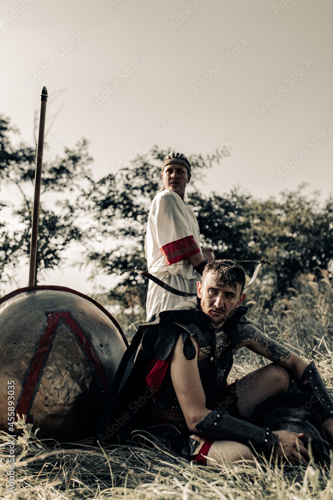 Ancient warrior and woman archer rest near shield and spear.