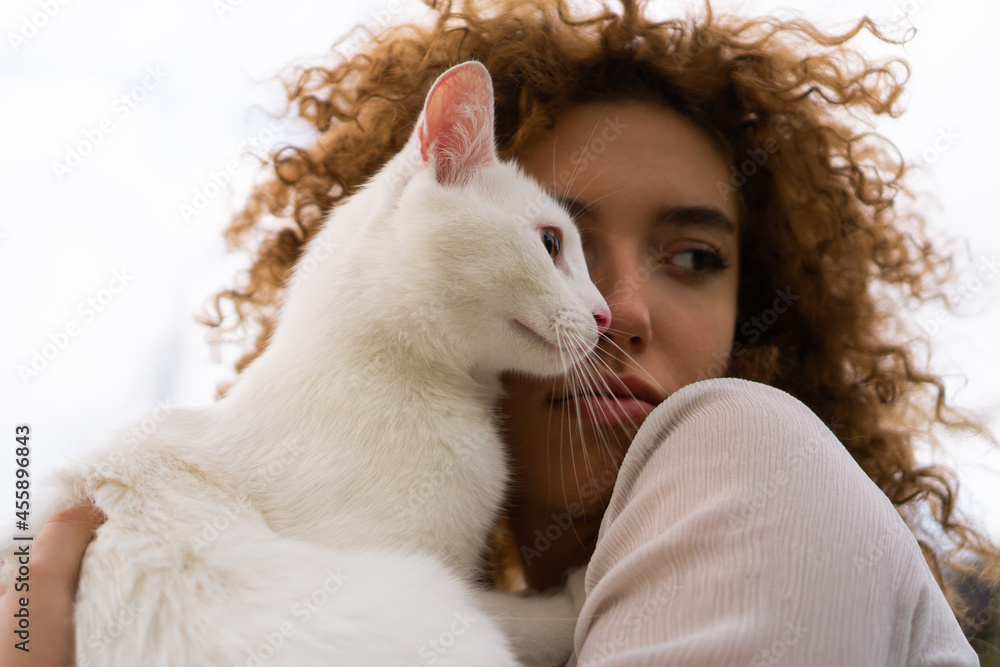 A beautiful teenage girl with curly hair holds a white cat close to her face. She is enjoying time with her pet.