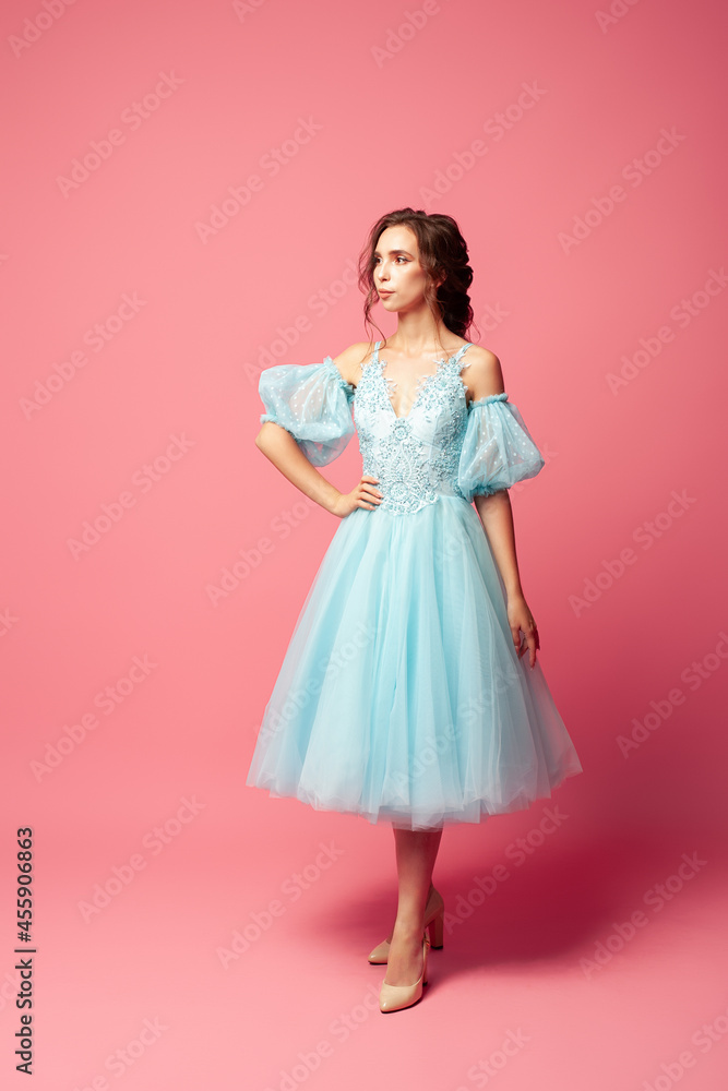 full-length portrait of elegant young woman in blue evening dress with full skirt and lantern sleeves, isolated on pink background. 