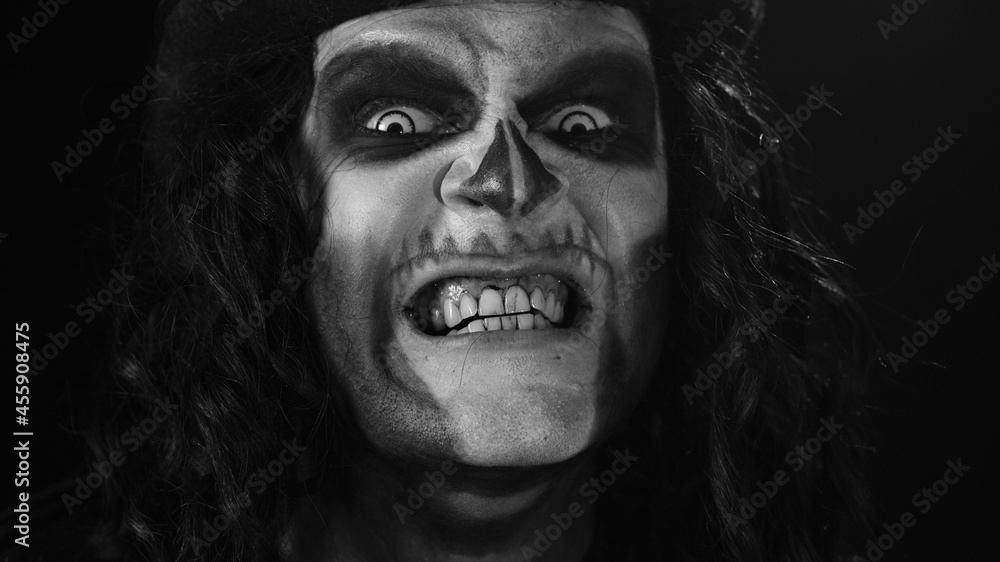 Close-up of frightening man face in skeleton Halloween cosplay. Crazy guy in creepy skull makeup looking at camera, showing dirty black teeth, trying to scare smiling. Dark background. Black and white