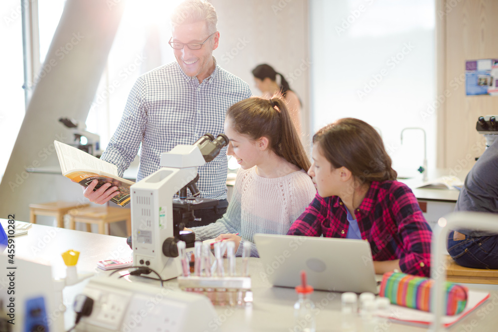 Obraz premium Male teacher helping girl students using microscope, conducting scientific experiment in laboratory classroom