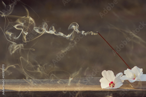 The incense stick was lit to perform spiritual practices. Smoke from incense and white flowers in a bowl on a wooden table. Background for design and advertising of meditation. Copyspace