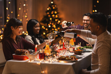 holidays, party and celebration concept - multiethnic group of happy friends having christmas dinner at home and drinking wine