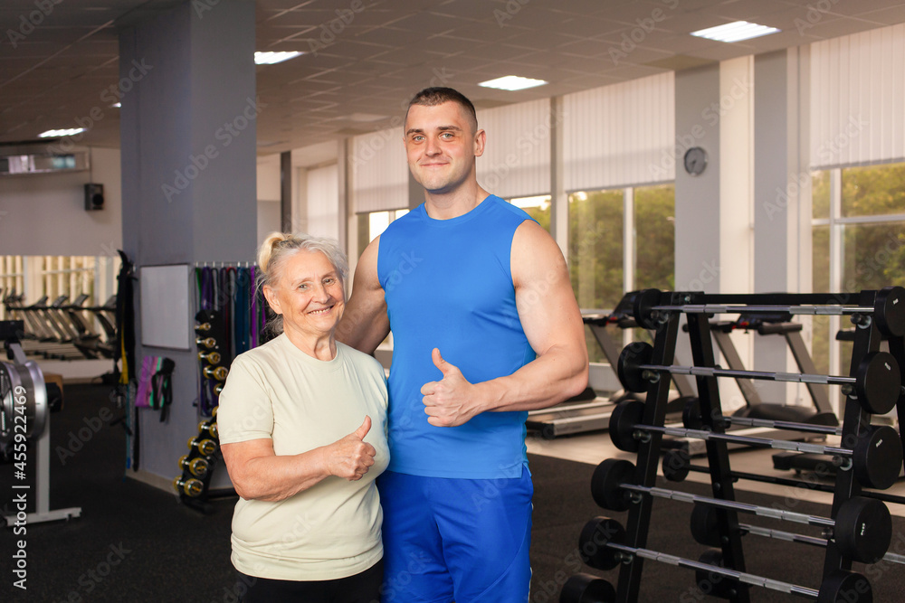 Obraz premium Portrait of a young coach in uniform and an old woman in gym. Physical culture for older people. Training and individual courses. Happy people smile, sport is cool, super, thumbs up. concept senior