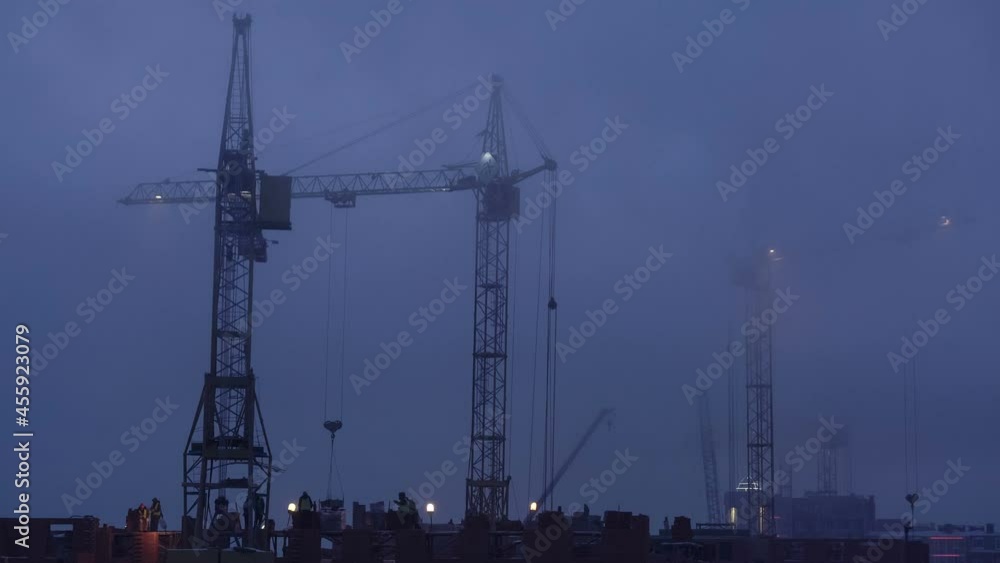 Construction site at dusk with purple sky. Silhouette of high tower cranes works on high-rise residential building site, lifts load. Construction site at dusk with purple sky. Silhouette of high tower cranes works on high-rise residential building site, lifts load.