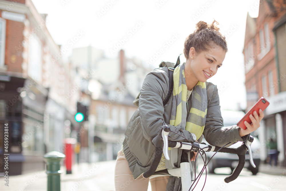 Young woman commuting on bicycle, texting with cell phone on sunny urban street