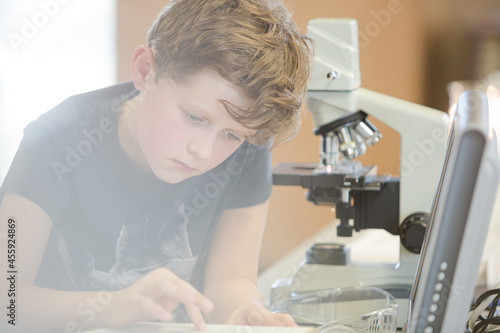 Fototapeta Naklejka Na Ścianę i Meble -  Boy student conducting scientific experiment at microscope and computer in laboratory classroom