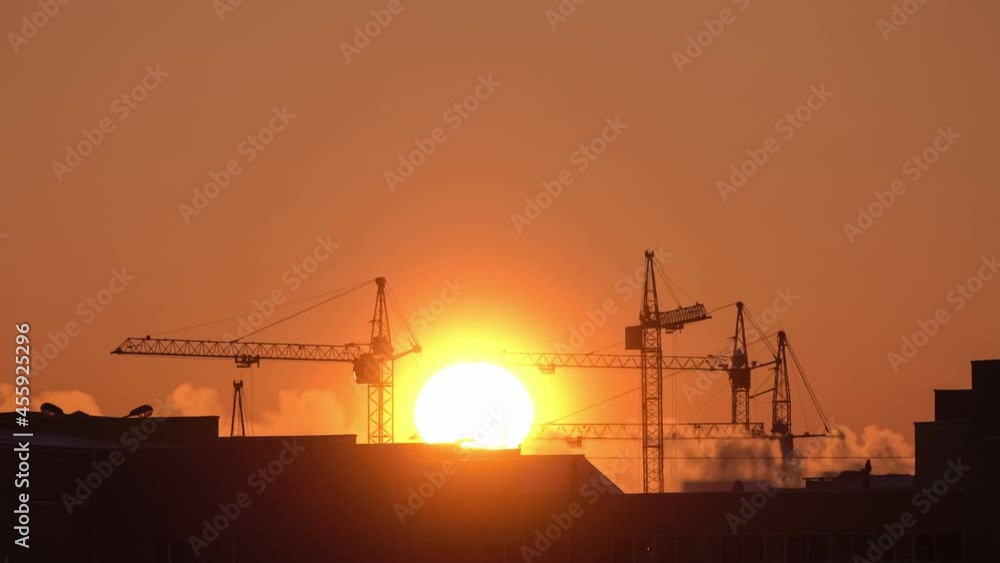 Construction site at orange sunset. Silhouette of high tower cranes works on high-rise residential building site, lifts load. Construction site at orange sunset. Silhouette of high tower cranes works on high-rise residential building site, lifts load.